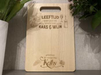 Houten snijplank met opschrift "leeftijd speelt geen rol, behalve voor kaas en wijn" en "gelukkige 50e verjaardag, Kathy" met bijhorende decoratieve elementen.