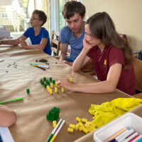 Kampdeelnemers rond een tafel; ze spelen een abstracte spellen met pionnen op spelborden die ze uittekenden op de rol papier die de tafel bedekt.